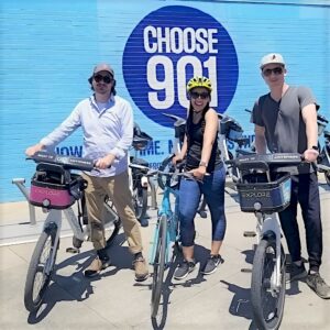 Three people standing with rental bikes in front of a blue wall that says "choose 901," showcasing a guide to Memphis biking.