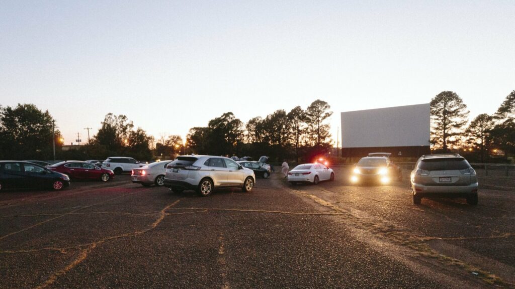 Cars parked at an outdoor drive-in movie theater in Memphis at dusk, with a large screen in the background and people walking around.
