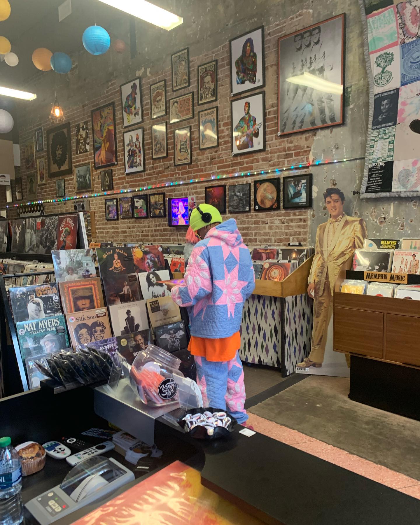 A person in a colorful costume inside a record store in Memphis, standing near posters and vinyl records, with a cardboard cutout of Elvis Presley in the background.