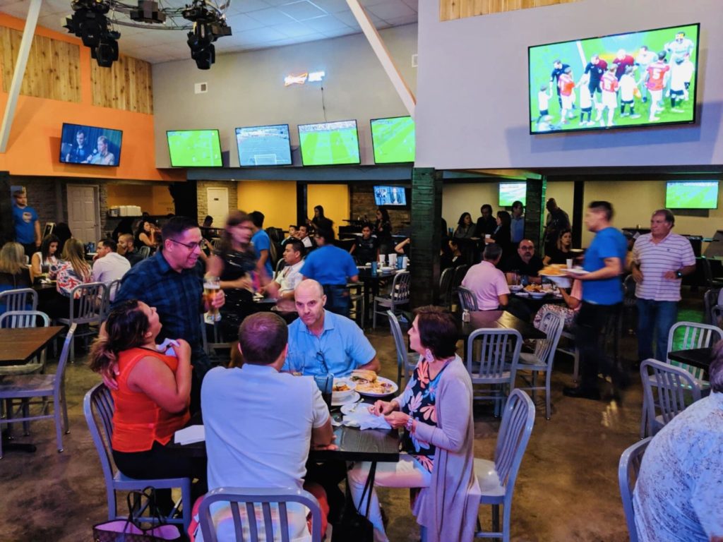 A group of people sitting at tables in a sports bar, watching a soccer match on the TV screens.