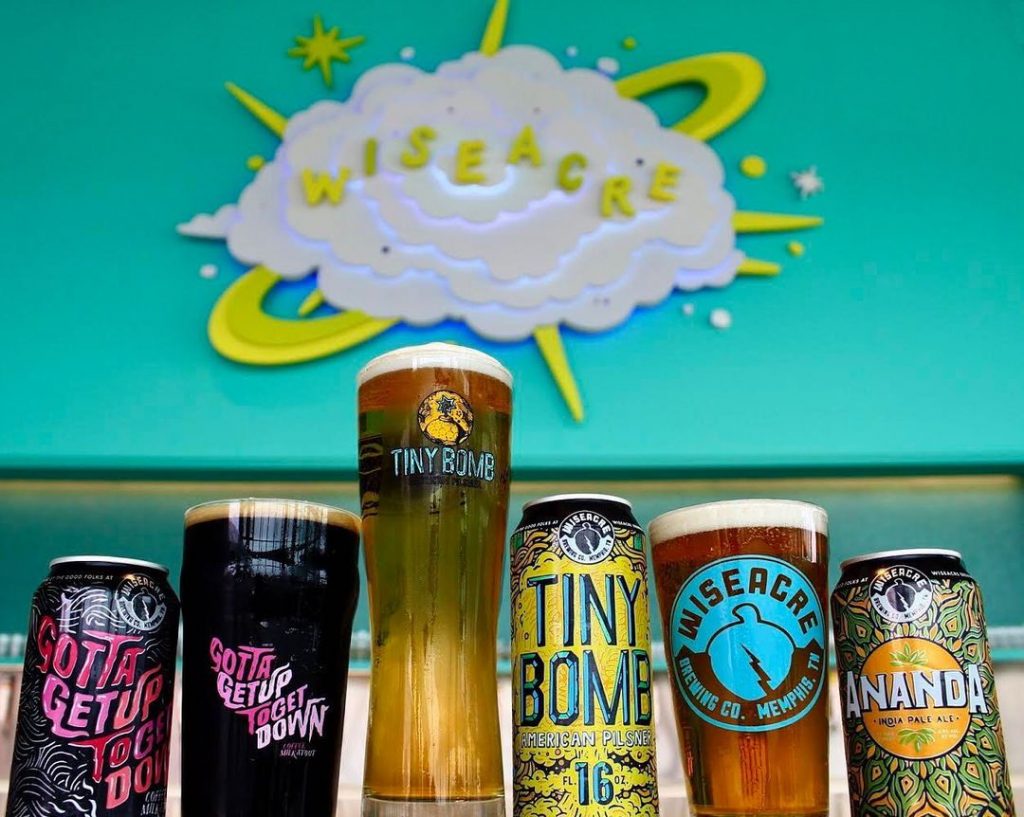 A lineup of beers from various breweries is displayed against a blue wall.