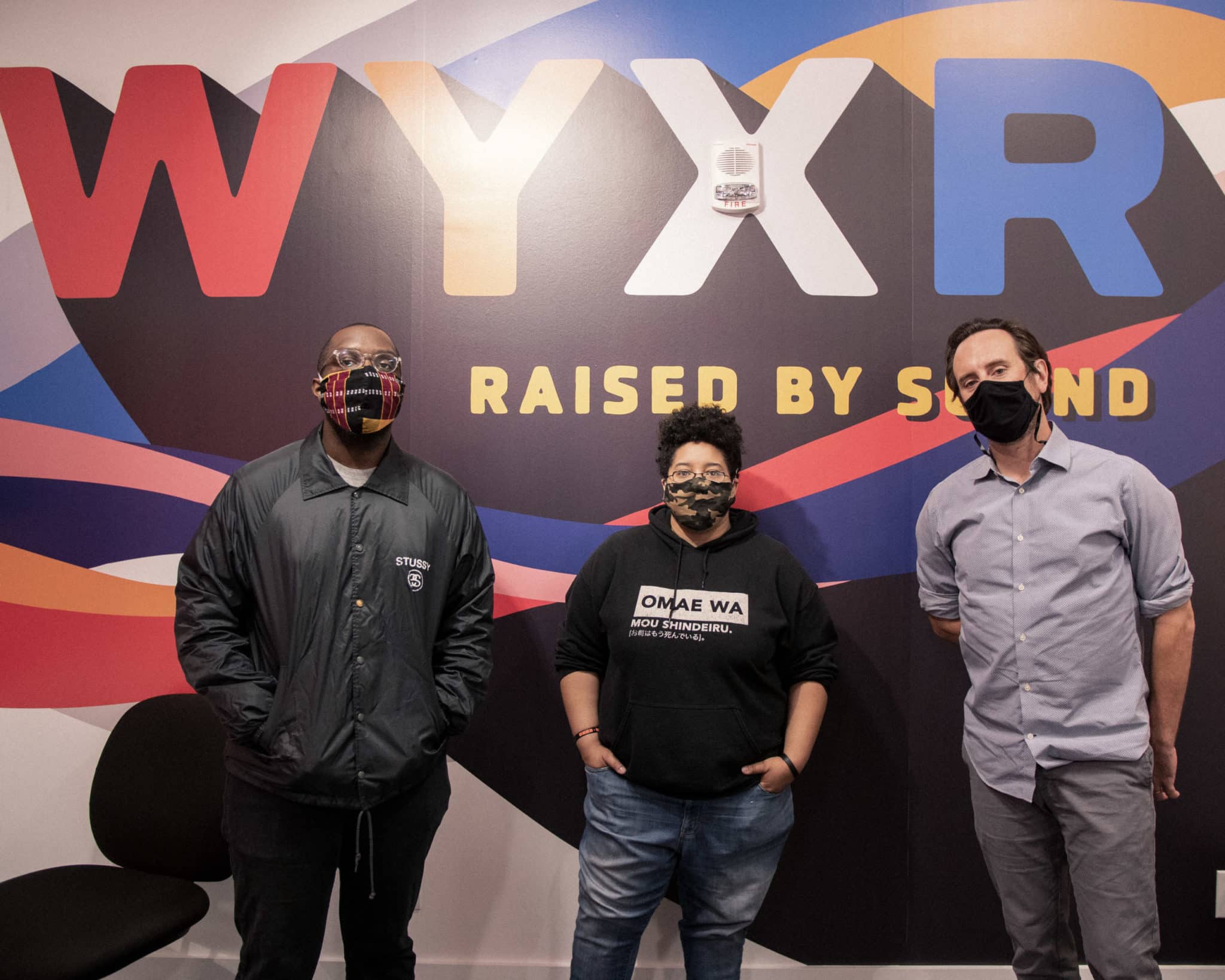 Three people standing in front of a wall with the word wyxr raised by sound.