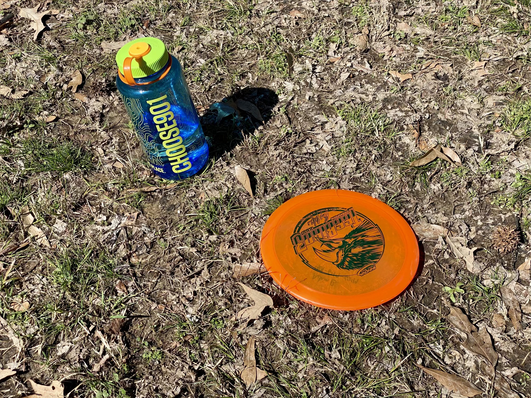 Orange disc and Blue Choose901 water bottle on ground at disc golf course in Memphis.