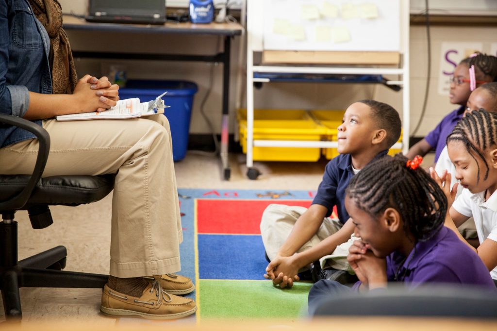 A teacher in a classroom with TFA Alumni sitting on the floor.