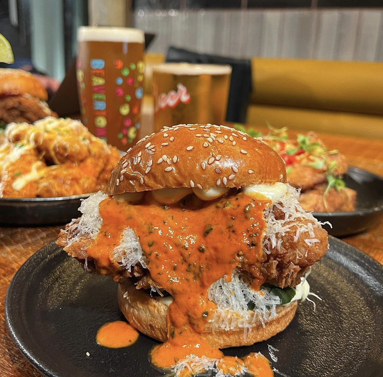 A late-night chicken sandwich with spicy sauce on a sesame bun, served with a beer and side dishes on a wooden table.