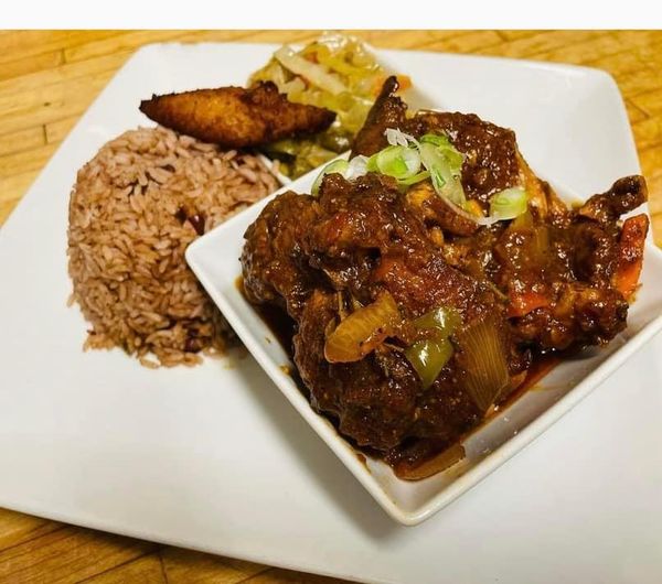 A plate of late night bites featuring Caribbean food with oxtail stew and onions in a white bowl, served with side dishes of rice and fried plantains.