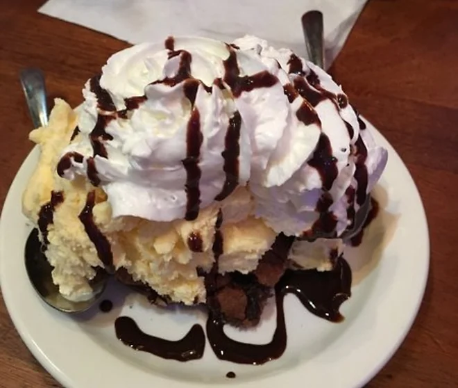 A large slice of pie topped with whipped cream and chocolate syrup, served on a white plate with forks on either side, perfect for late night bites.