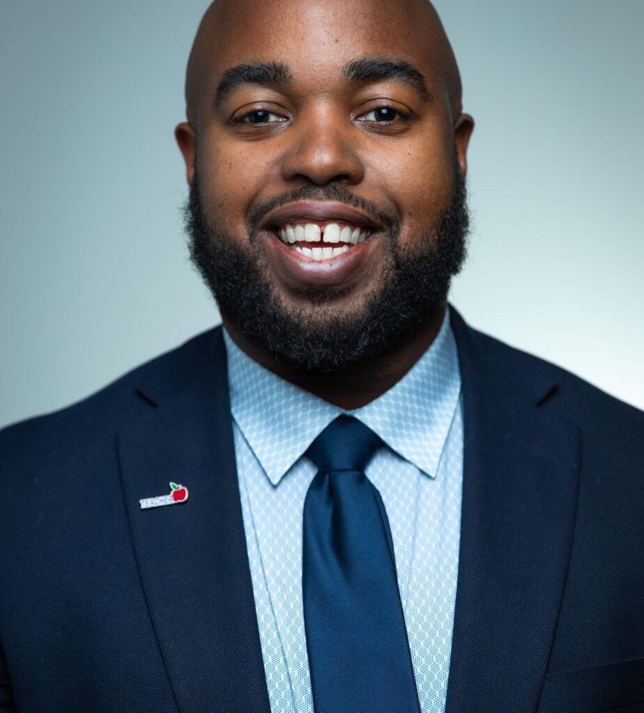 A black man in a suit and tie smiling while representing Teach For America.