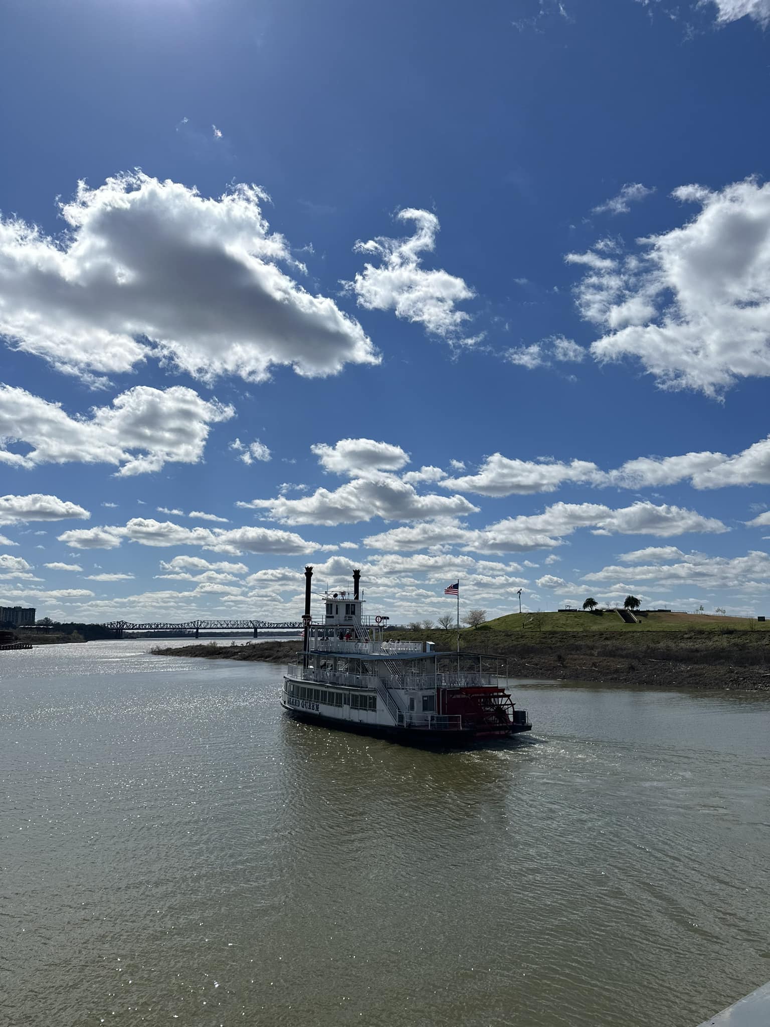 A paddle steamer on the Memphis river under a partly cloudy sky, with a bridge in the background and green riverbanks.