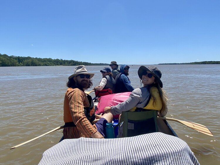 Group of people in a boat on the Memphis river, some wearing hats and sunglasses, sitting with paddles, looking back and smiling under a clear blue sky.