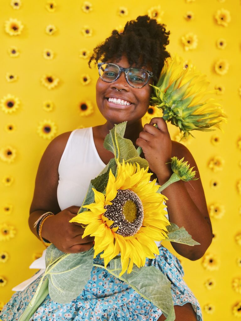 A woman holding sunflowers in front of a yellow wall, symbolizing rising above with Clover Boggan.