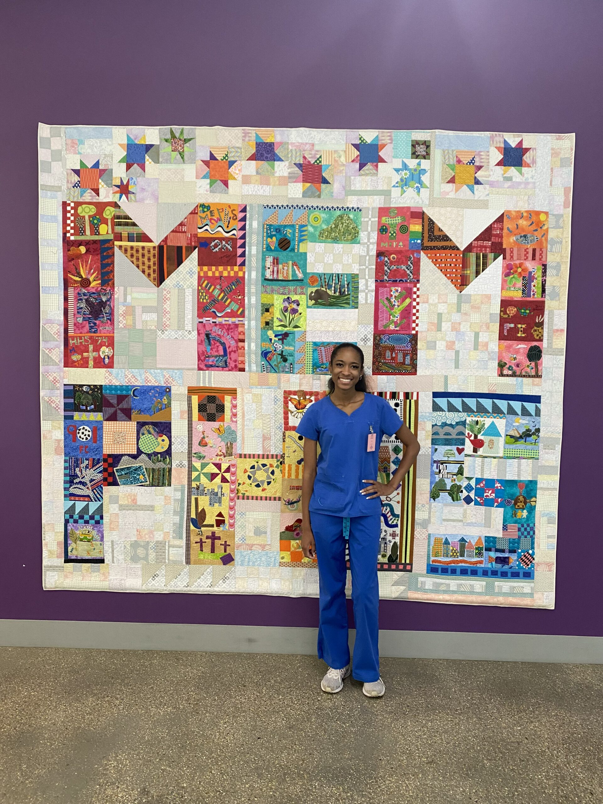 A nurse in scrubs standing in front of a quilt at NexGen Haven Newson.