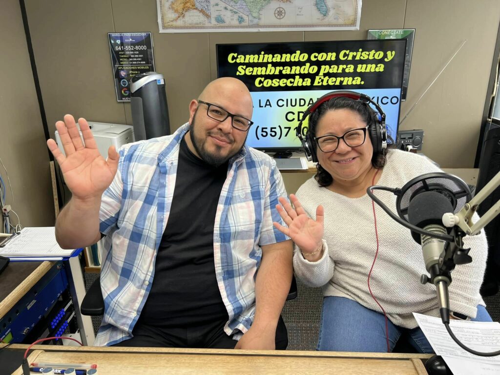 Two people smiling and waving at the camera from inside a radio studio with visible microphones and a Spanish language banner promoting the Latinx community in Memphis in the background.