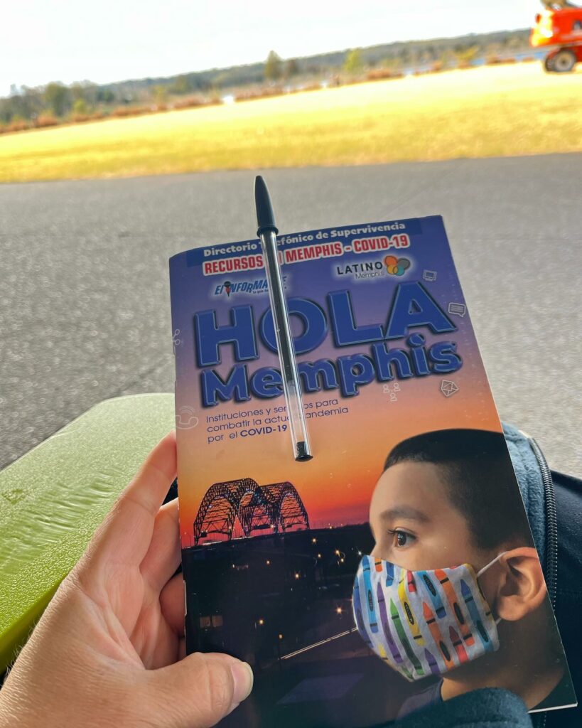 A person holds a Spanish-language magazine titled "Hola Memphis" featuring resources for COVID-19, serving the Latinx community in Memphis, with a child in a mask sitting next to them on a bus