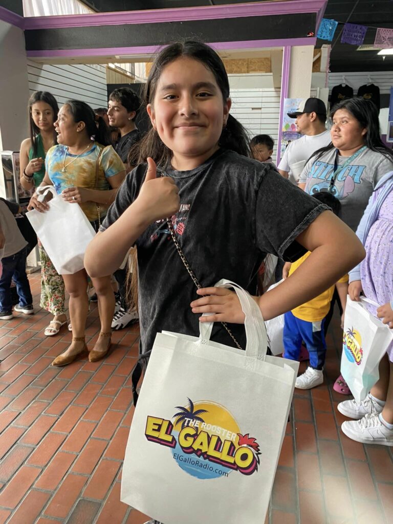 A smiling girl wearing a black t-shirt holds a tote bag with "el gallo radio" logo, representing the Latinx community in Memphis, and gives a thumbs up in a busy market area.