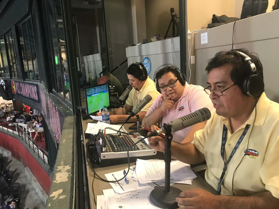 Three broadcasters from the Latinx community in Memphis commentate on a soccer match from a press box overlooking the stadium, using microphones and observing the game on a screen.