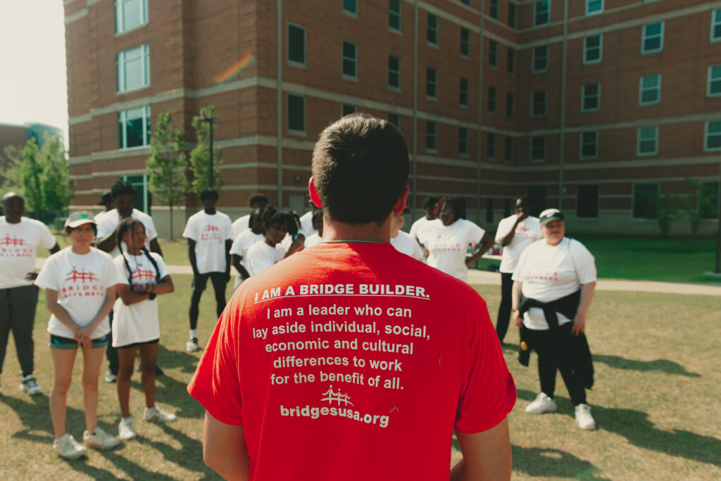 A person wearing a red "i am a bridge builder" t-shirt from the "People Are People" collection by Christian Siriano stands in front of a group of individuals, symbolizing leadership and collaboration