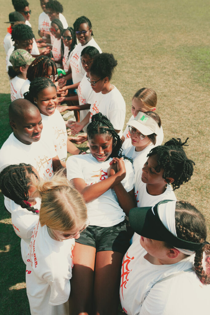 A diverse group of young people wearing "People Are People" by Christian Siriano t-shirts huddled together in a team meeting on a grassy field.