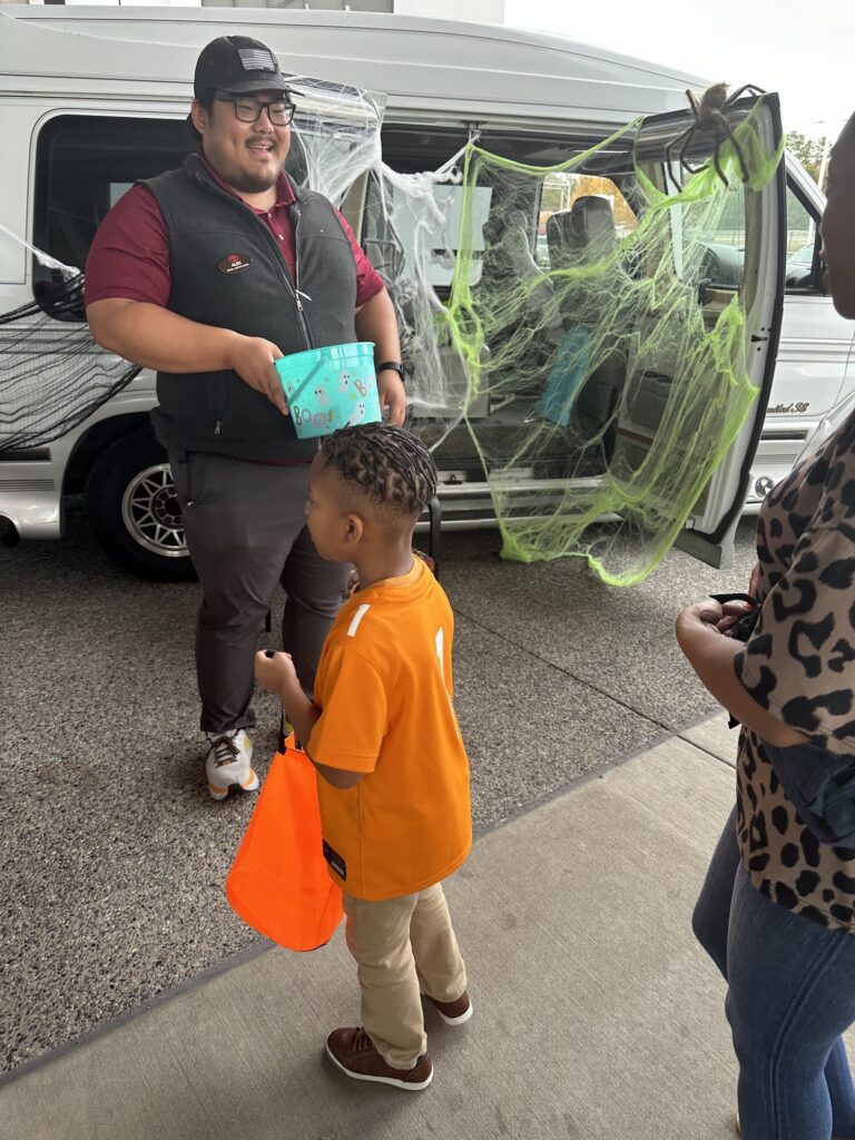 A young child in an orange shirt and brown pants receives a teal-colored box of School Supplies from a smiling person standing next to a van decorated with green cobwebs for a festive occasion.