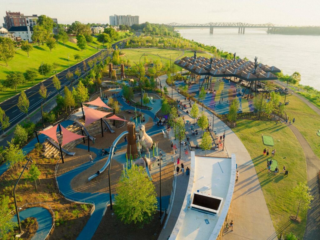An aerial view of a riverside park with a playground, walking paths, and green spaces during the Riverbeat Music Festival, with people engaged in various recreational activities.