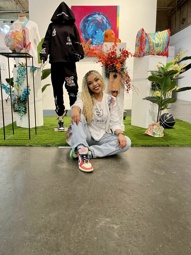 A person sitting on the floor in an art gallery area with colorful artwork and mushroom-themed floral arrangements in the background.
