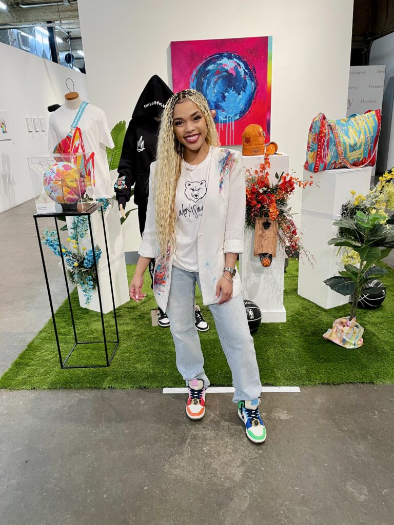 A smiling person standing in an art space with colorful paintings, mushroom sculptures, and flower arrangements around.