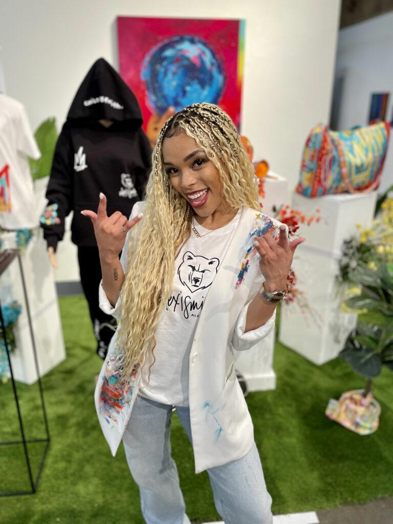 A person posing with a peace sign in an art gallery surrounded by clothing items, mushrooms, and artwork on display.