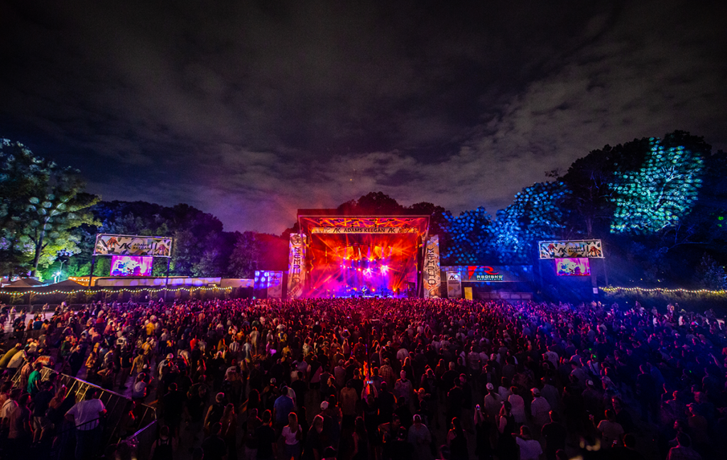 A large crowd of spectators gathered at the Riverbeat Music Festival during nighttime, with a brightly lit stage in the foreground and trees illuminated with blue lights in the background.