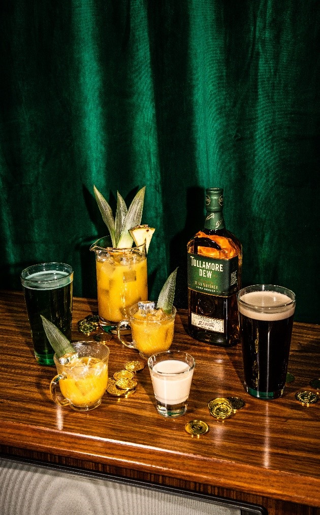 A selection of alcoholic beverages on a wood surface against a dark green backdrop, including cocktails with pineapple garnish, a bottle of Tullamore Dew whiskey, and a dark stout beer at St.