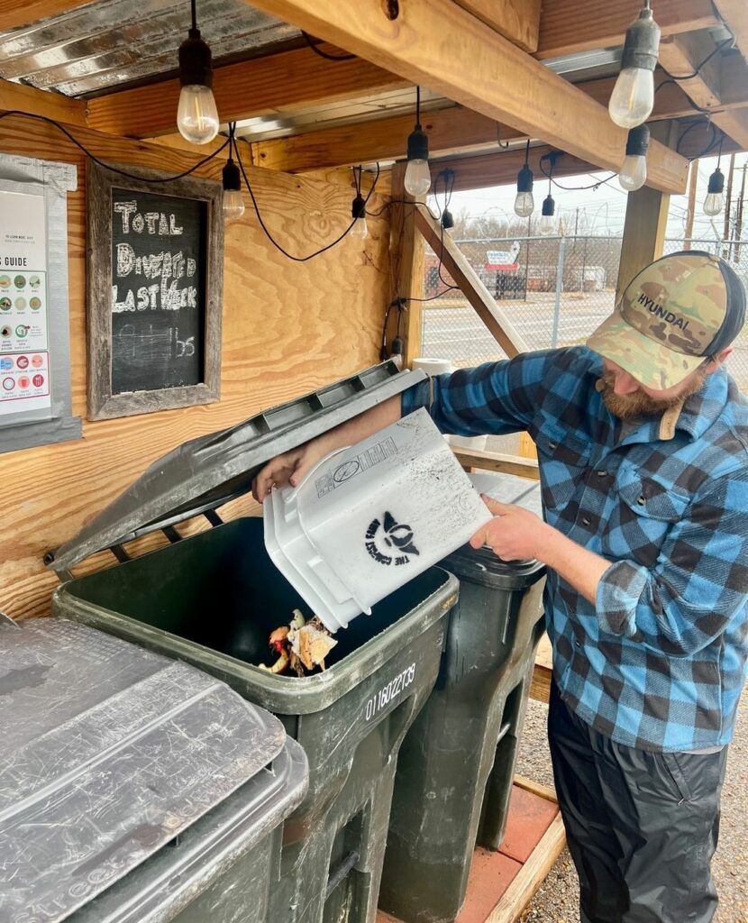 A man in a plaid shirt and cap pouring contents from a bucket into a compost bin at a sustainable recycling station.
