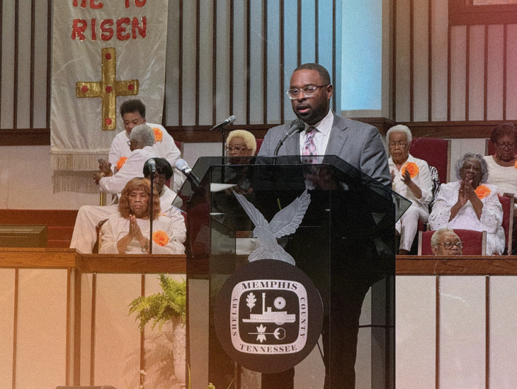 Mayor Paul Young delivers a speech at a church podium with the congregation seated behind him, in a setting with religious symbols and the text "he is risen" visible.