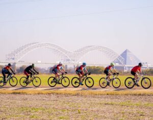 A group of cyclists enjoying Memphis biking on a dirt track in the foreground with a large steel arch bridge in the distant background.