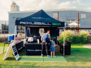 A vendor booth labeled "Getting Started with Pickleball 901" in Memphis, with two customers, one adult and one child, looking at merchandise outside under a tent.