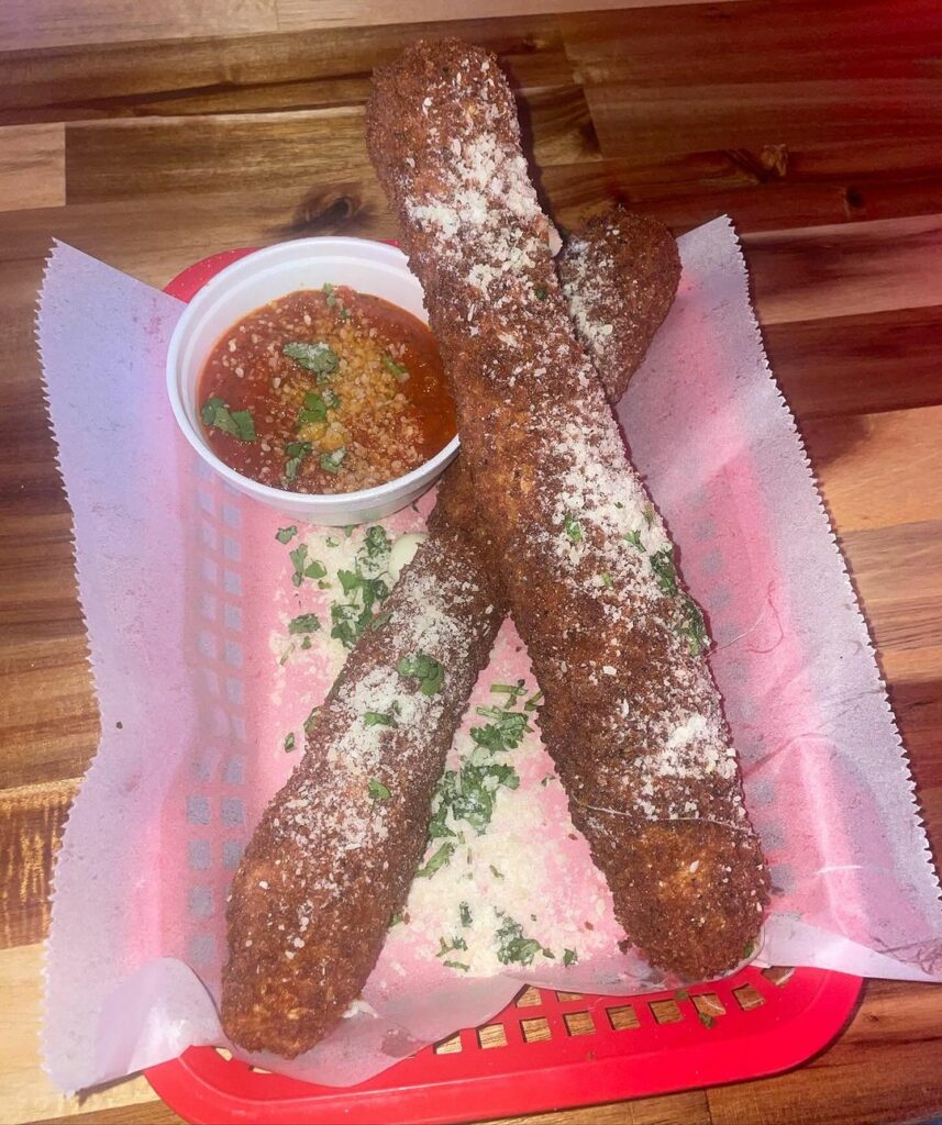 Two large mozzarella sticks covered in parmesan cheese and herbs, served with marinara sauce in a red basket on a wooden table, perfect for late night bites.