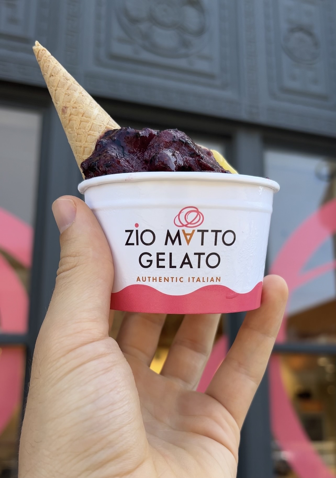 Hand holding a cup of Zio Matto Gelato with a waffle cone inserted. The gelato appears to be a dark berry flavor. The background shows a store facade, making it a must-try from the Ultimate Guide to Frozen Treats in Memphis.