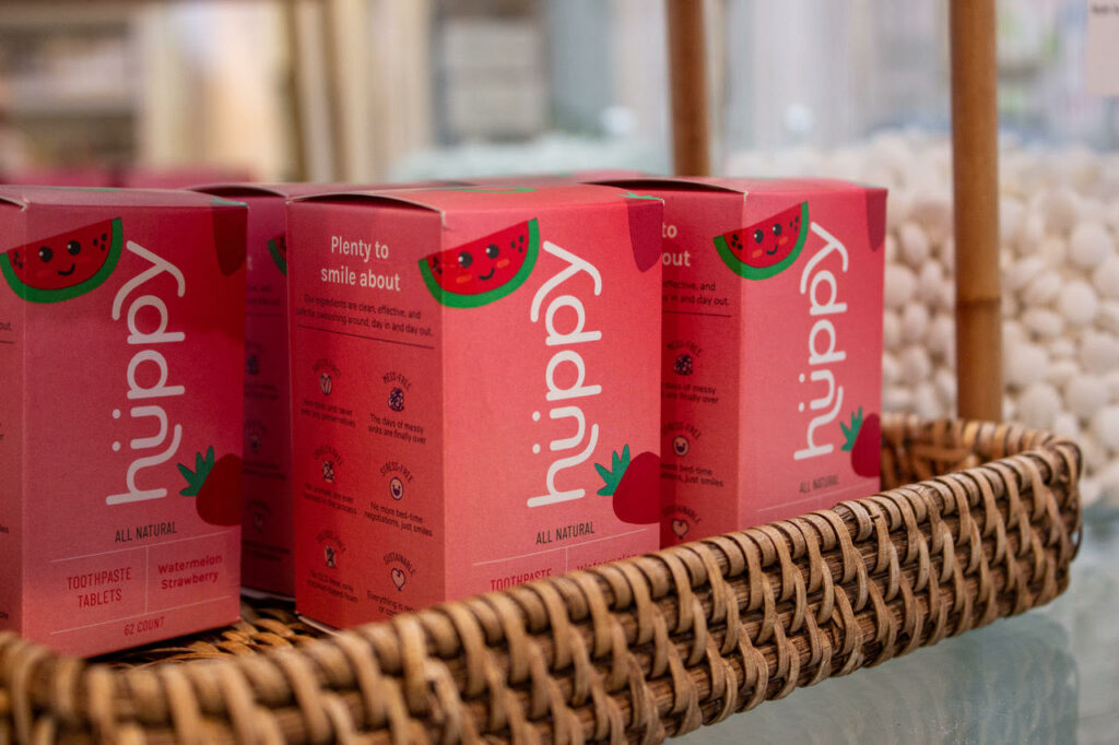 Rows of red toothpaste boxes labeled "huppy" in a sustainable wicker basket on a store shelf, featuring watermelon graphics and text promoting natural ingredients.