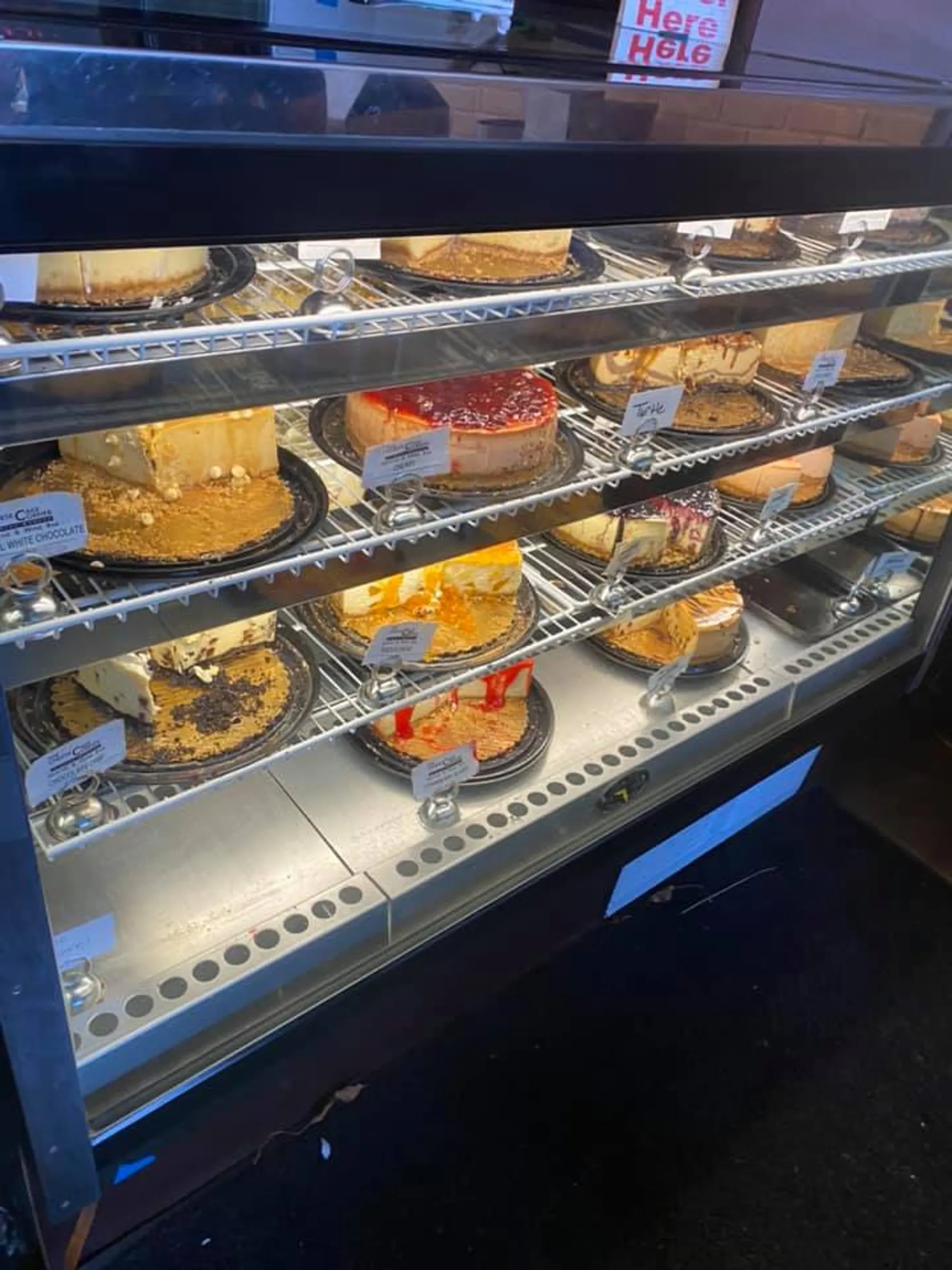 Assorted cakes on display in a bakery case, including cheesecakes and layered cakes, ideal for late night bites, with price tags visible.