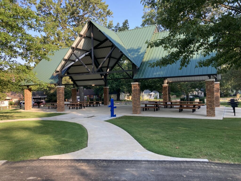 A Memphis Parks pavilion with a large green roof, brick columns, picnic tables, and a concrete pathway leading to it, surrounded by trees.