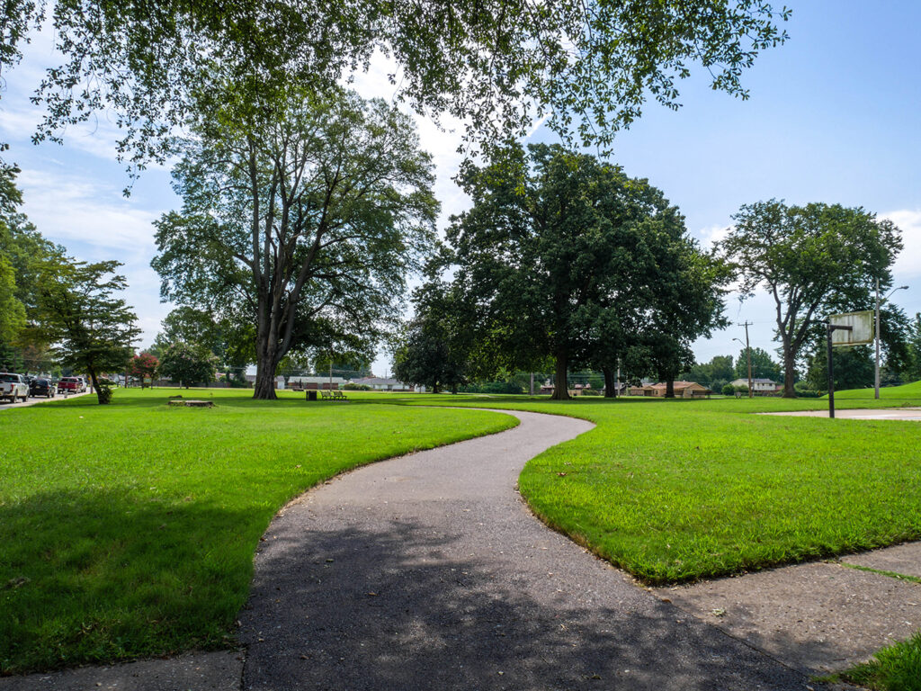 A paved path winding through a lush Memphis park with tall trees, grassy areas, and a basketball court under a clear sky.