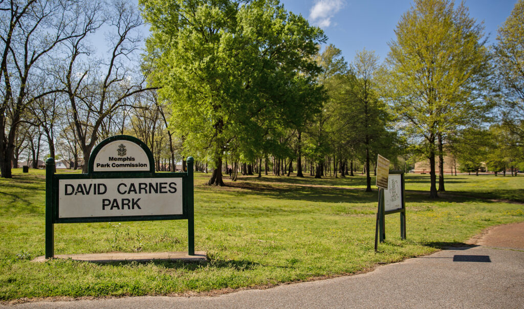 Entrance of David Carnes Park, featuring a green sign with the park name, managed by Memphis Parks Commission, with trees and a clear sky in the background.