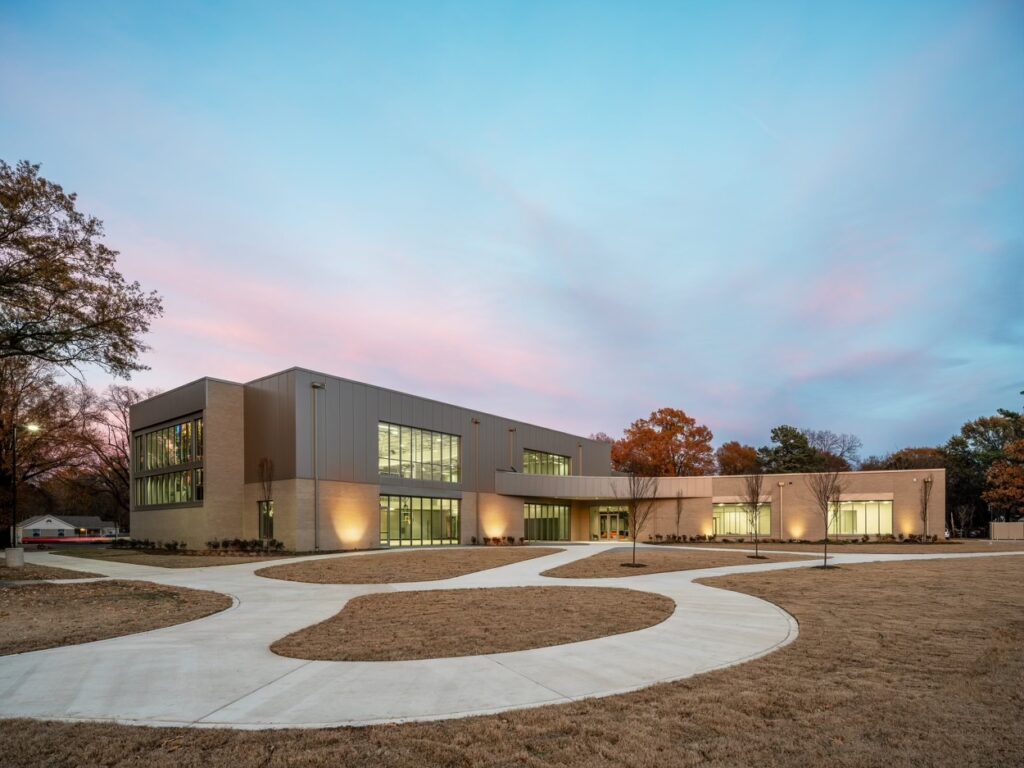 Modern educational building with large windows illuminated at twilight, foreground showing a curved pathway in Memphis Parks on a grassy area under a pastel sky.