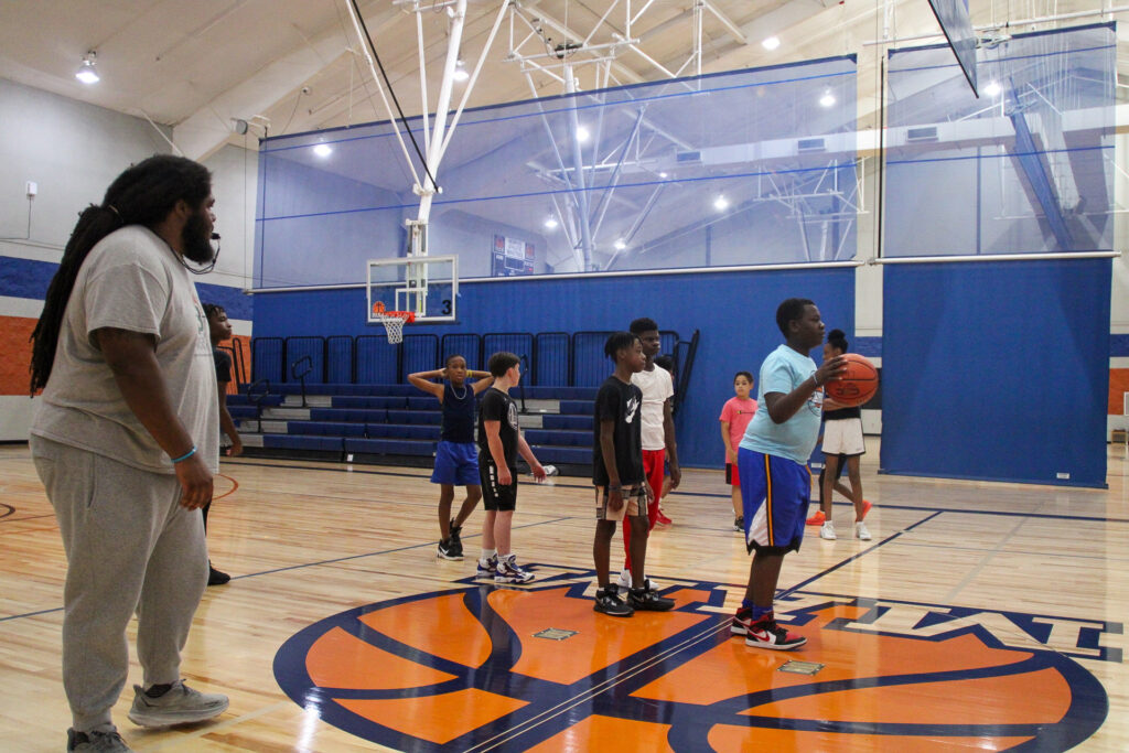 A coach observes young players during a basketball practice in an indoor gymnasium as part of summer programs.
