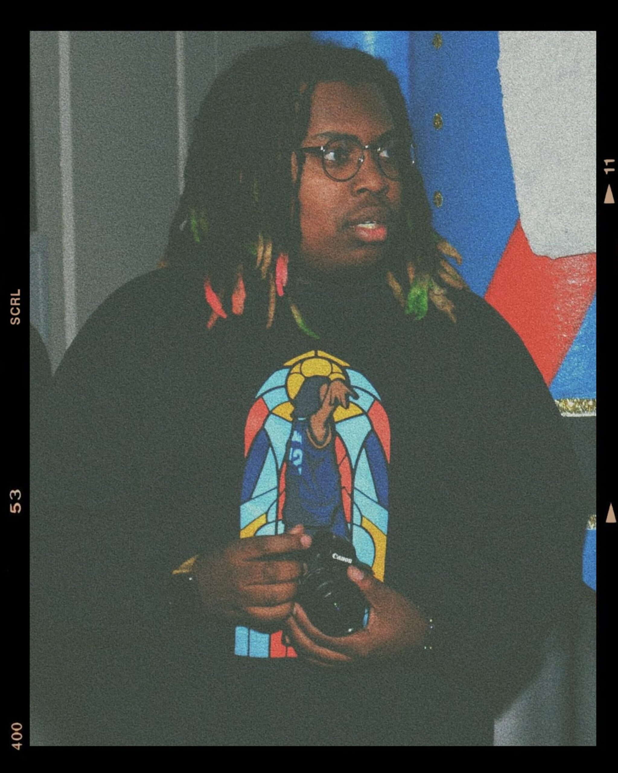 Young man with dreadlocks holding a camera, standing in front of a colorful abstract painting at the Riverbeat Festival, photographed with a vintage filter.