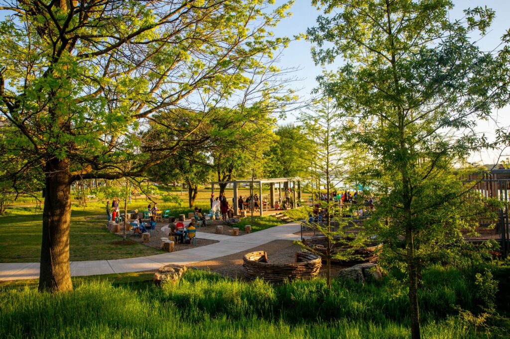 A vibrant Memphis park scene with people enjoying a sunny day near a pergola, surrounded by abundant green trees and paved pathways.