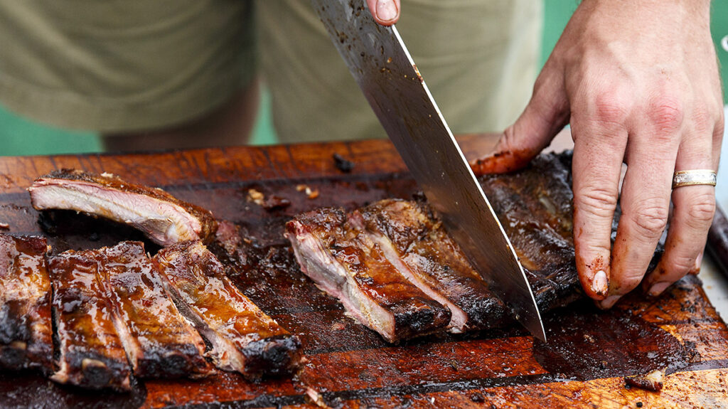 A person slicing barbecued ribs on a wooden board with a large knife at the Memphis BBQ Fest.