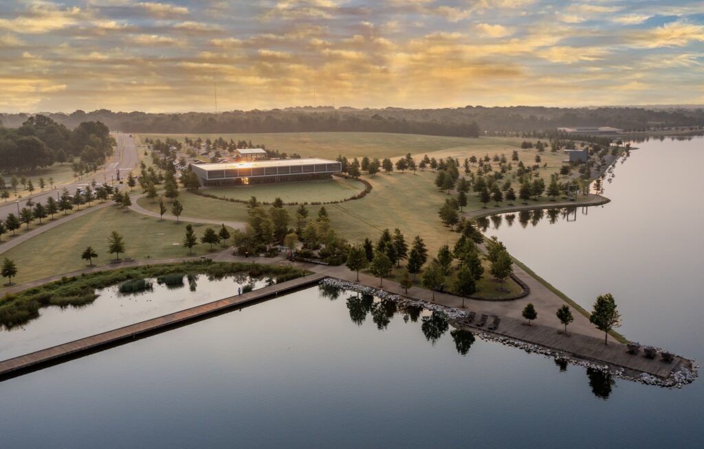 Aerial view of a modern building beside a lake in Memphis Parks with a surrounding park and parking area at sunrise, reflecting clouds in the water.