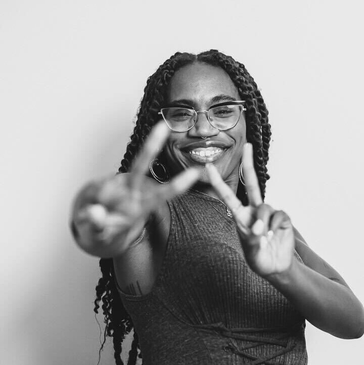 A joyful woman with braids and glasses making a peace sign with her hand at the Riverbeat Festival, against a plain background.