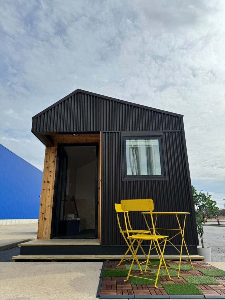 Small, modern black cabin with a slanted roof and open door, featuring a yellow chair and table set on a wooden deck, against a blue sky at the hospitality hub.