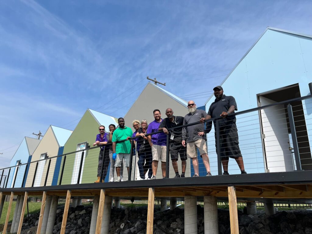 Group of seven diverse adults smiling and posing on a wooden deck in front of triangular-roofed houses under a clear blue sky, showcasing Memphis's hospitality hub.